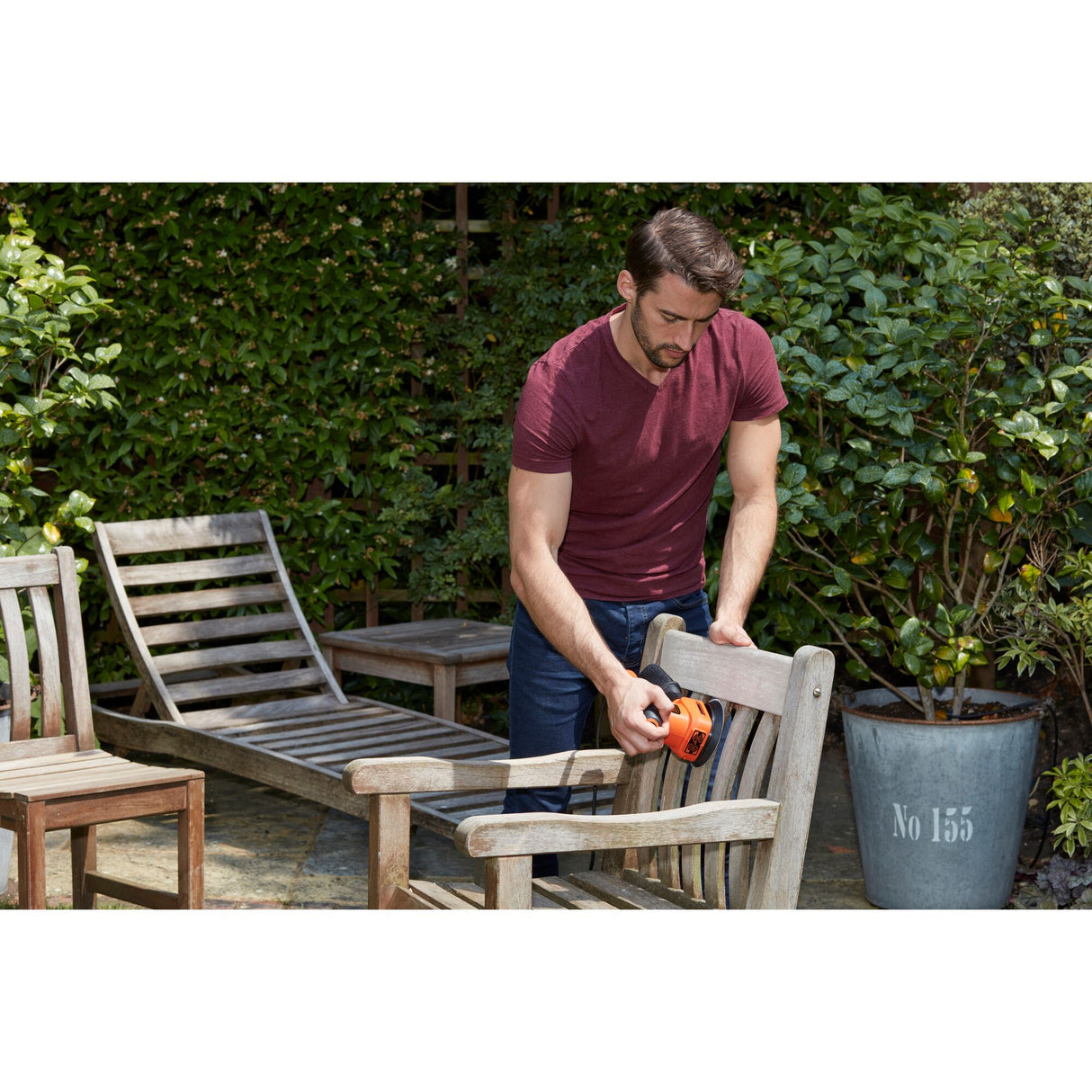 Homme utilisant une ponceuse Black+Decker orange pour restaurer une chaise en bois dans un jardin.