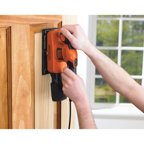 Ponceuse électrique Black+Decker orange utilisée sur porte en bois clair.